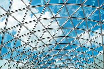 Glass ceiling with geometric pattern reflecting a vibrant blue sky and scattered clouds. The architectural design creates a modern, airy, and open ambiance in the space.