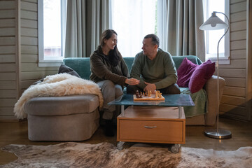 Mature couple sitting on sofa at home and spending time together playing chess, talking and relaxing in cozy living room interior. Home Leisure Activities