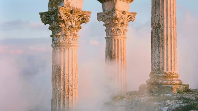 Ancient stone columns rising through soft clouds at sunrise with detailed corinthian capitals and weathered classical architecture