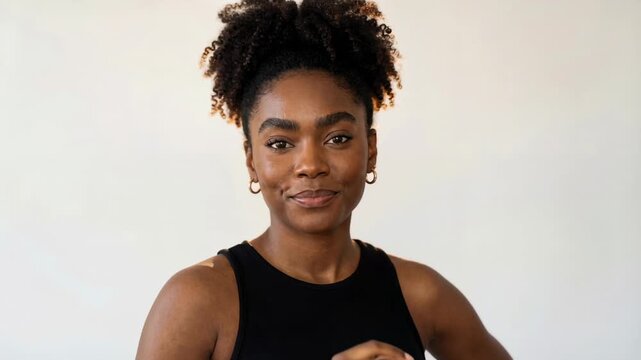 Confident young woman with natural curly hair posing in black sleeveless top against soft neutral background indoors