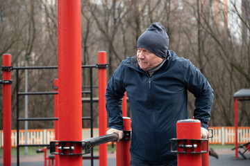 Adult man exercise on parallel bars in an urban outdoor gym after rain. Dressed warmly, concentrating on fitness routine amidst city backdrop, self discipline. Daily routine, all season.