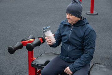 Adult man sits on an outdoor gym bench holding protein shake, wearing casual vest. He relaxed, enjoying moment of rest during workout.