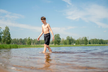 Young boy walking through shallow water at a lake on a sunny day, creating small ripples with each step. Surrounded by nature and trees under a clear sky. © Наталья Майшева