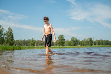 Young boy walking through shallow water at a lake on a sunny day, creating small ripples with each step. Surrounded by nature and trees under a clear sky.