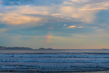  p&ocirc;r-do-sol na Praia Palmas do Arvoredo cidade de Governador Celso Ramos Santa Catarina Brasil regi&atilde;o da Grande Florianopolis ao fundo um arco-&iacute;ris na costa de Bombinhas Mariscal