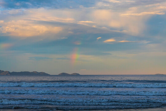  p&ocirc;r-do-sol na Praia Palmas do Arvoredo cidade de Governador Celso Ramos Santa Catarina Brasil regi&atilde;o da Grande Florianopolis ao fundo um arco-&iacute;ris na costa de Bombinhas Mariscal