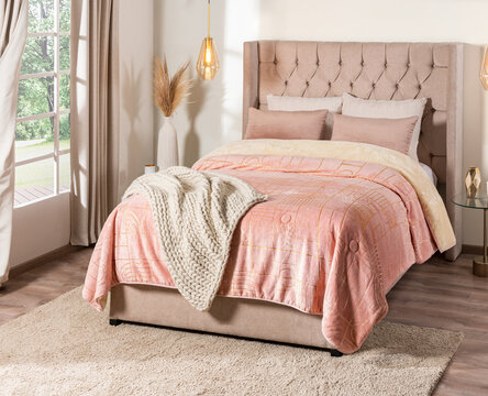 Scandinavian-style bedroom showcasing a upholstered bed, geometric patterned quilt, draped blanket, a glass side table with brass accents, ceramic vase holding dried pampas grass, lighting window.