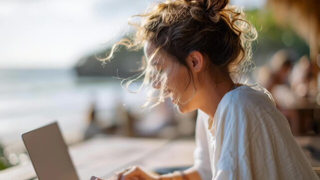 A woman is sitting on a beach with a laptop in front of her. She is focused on her work, possibly typing or browsing the internet. The beach setting suggests a relaxed and leisurely atmosphere