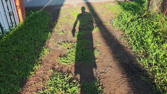 The setting sun casts a long shadow of a man walking along a grassy path that eventually meets a sidewalk. The scene is captured from a first person perspective