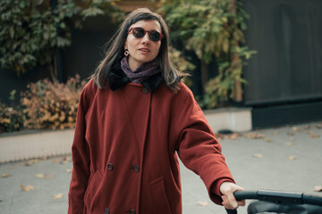 Modern adult mother in red coat have walk with child in pram in city