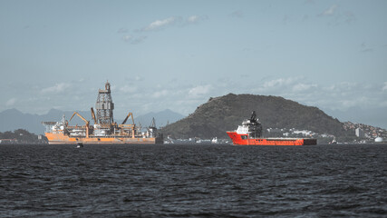 Offshore drilling rig and red supply vessel on a coastal bay, industrial oil and gas operation with cranes and derrick, distant shoreline city and mountain under wide blue sky