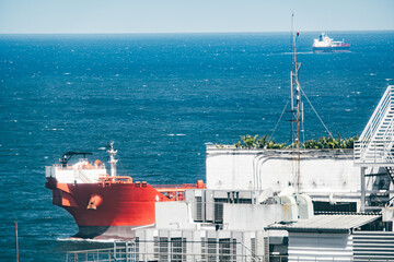 Telephoto view of red cargo tanker passing an industrial harbor, blue ocean with whitecaps, rooftop equipment and antenna mast in foreground, another ship on distant horizon