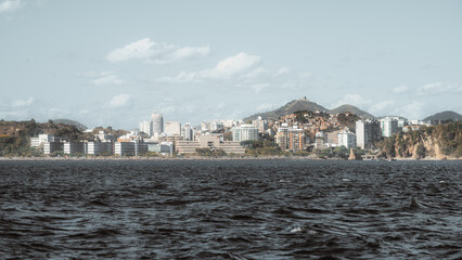 Telephoto view of Niteroi skyline across Guanabara Bay, Brazil, with choppy dark water foreground, modern building facade and hillside neighborhood under soft cloud and blue sky