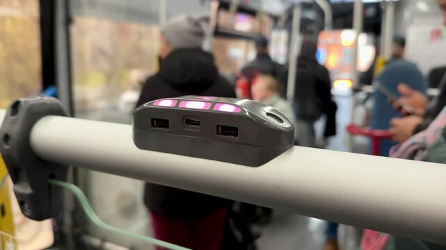 Usb Charging Hub Mounted On Handrail, Pink Led Indicators Glowing, Three Usb Ports And Compact Display, Blurred Passenger Silhouette In Background, Bus Interior During Commute, CloseUp