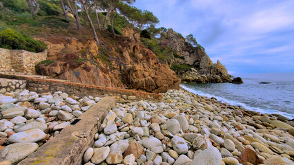 Rocky Mediterranean coastline with pine trees on cliffs and a pebble beach under a cloudy sky.