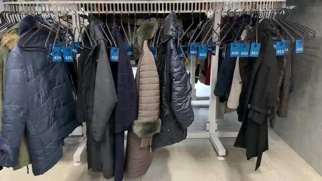 Rows Of Winter Coats Hanging, Shop Owner Sorting Preloved Inventory Beneath Crowded Rack, Mixed Textures Wool Denim Leather, Muted And Bright Colors, Cozy Cluttered Atmosphere, Sustainable