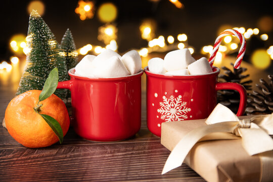 Two red cups of cocoa with marshmallows, a tangerine, a gift box. Festive lights, New Year, Christmas.