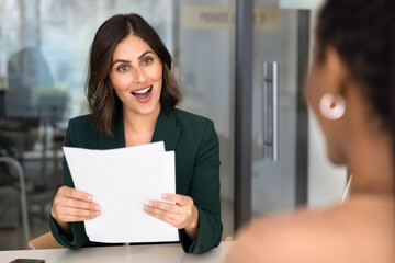 Obraz premium Excited positive HR manager woman meeting with job applicant at office table, holding paper documents, resume, learning about spectacular professional skills of candidate, gasping in surprise