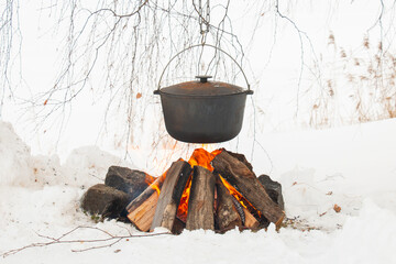 a kettle with hot tea brewed in it hanging over the fire. On a hook on a tripod, winter cooking outdoors.