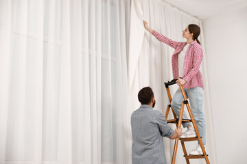 Couple with ladder hanging up window curtains at home, low angle view. Space for text