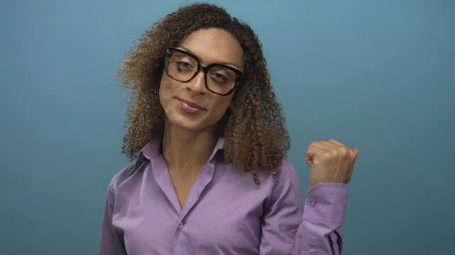 Transgender woman wearing glasses points thumb in studio with blue backdrop and smiles; confidence.