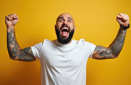Bald man with beard and tattoos raises fists in air, shouting loudly with joy and excitement. He celebrates a win against a bright yellow studio backdrop. Success achieved.