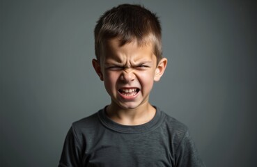 Fototapeta premium Young boy scrunches face in studio. Child shows anger grimace, brows furrowed tight. Kid expresses frustration, frustration, defiance, maybe pain. Isolated portrait. Youngster unhappy, upset, mad.