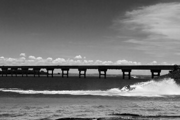 Black and white photo of jet ski crossing water beneath long bridge under dramatic sky 
