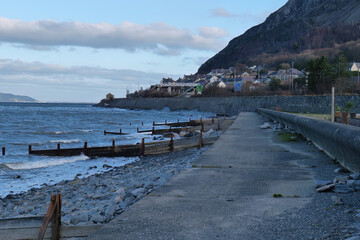 Coastal path stretches alongside a turbulent sea, backed by a mountain village under a cloudy sky; cool blues, grays, and muted light in Llanfairfechan - Wales - UK