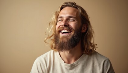 Handsome bearded man with long blonde hair laughs joyfully. Studio portrait shows positive emotion, natural smile. Male model wears casual shirt on plain background. Happy expression conveys genuine