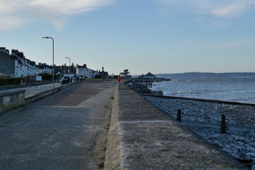 Coastal road stretches alongside a pebble beach, lined with houses under a bright sky, creating a serene, distant, and slightly muted atmosphere in Llanfairfechan - Wales - UK

