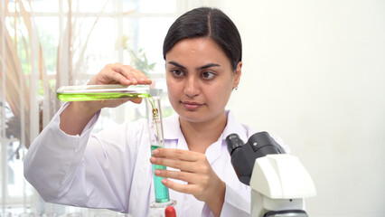 Focused female scientist pouring green liquid from test tube into graduated cylinder in laboratory. Young researcher conducting chemistry experiment or analyzing plant extract concept.
