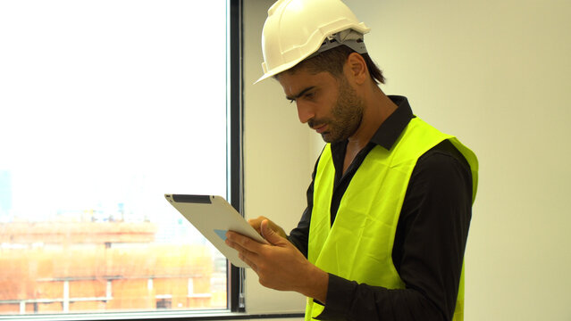 Professional Middle Eastern engineer wearing safety vest and hard hat using digital tablet near window. Focused construction manager checking project plans with building site background. - Powered by Adobe