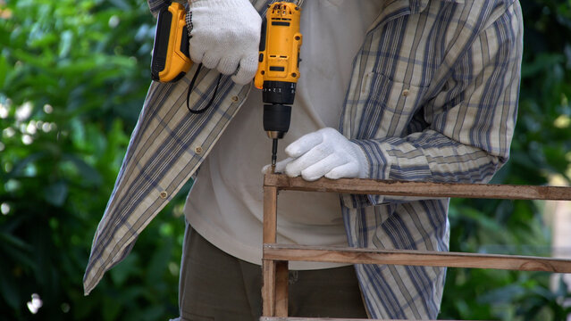 Close up carpenter hands wearing gloves using yellow cordless electric drill to screw into wooden frame. DIY handyman repairing furniture in garden workshop renovation concept. - Powered by Adobe