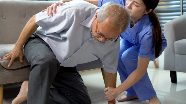 Professional Asian female nurse or caregiver helping elderly man stand up from floor at home. Young medical staff supporting senior patient suffering from pain or fall accident concept.