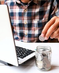 man's hand putting coins into a jar while working on a laptop