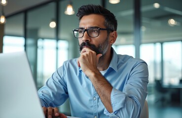 Man in glasses ponders project at desk with laptop. Indian pro thinks deeply, considers options for startup growth in modern office setting. Focused businessperson plans strategy. Digital world.