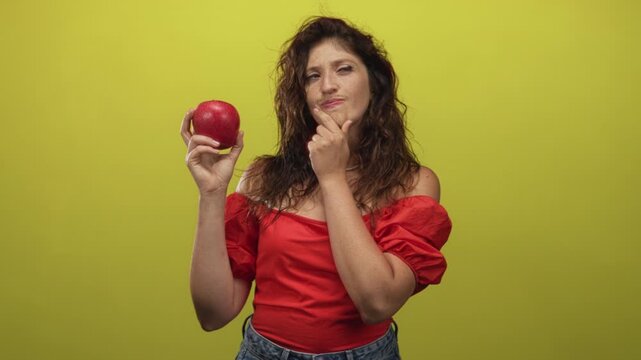 Young hispanic woman holding red apple and hand on chin in studio with yellow background, wearing red off shoulder top and denim jeans; curiosity choice.