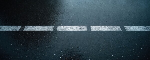 Wet asphalt road surface with white lane markings. Droplets create texture on dark grey pavement. Horizontal perspective shows sparse lines for traffic guidance.