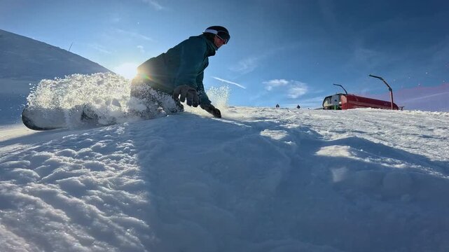 LENS FLARE, CLOSE UP, LOW ANGLE VIEW, SLOW MOTION: Male rider falls forward while snowboarding on ski slope, his hands digging into snow and blasting powder toward camera on sunny winter day in Alps.