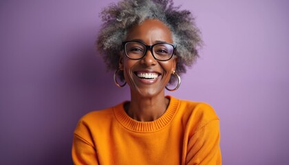 Obraz premium Elderly black woman with gray curly hair wears glasses and an orange sweater, smiling happily indoors against a purple backdrop. Her expression radiates warmth and contentment.