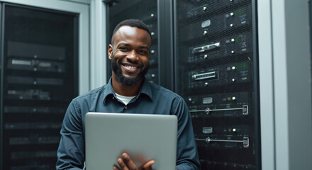 Smiling Black man holds laptop in server room. Technician works with computer hardware, network racks, data center equipment. pro manages systems, checks connections, maintains tech infrastructure.