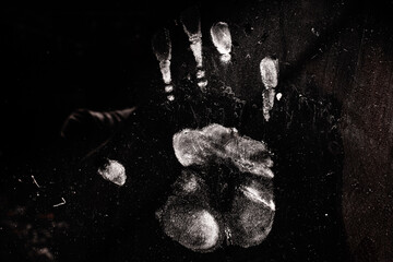 A white palm print contrasts against a pitch black background. The surface appears dusty and scratched, creating a mysterious, eerie, or forensic atmosphere.