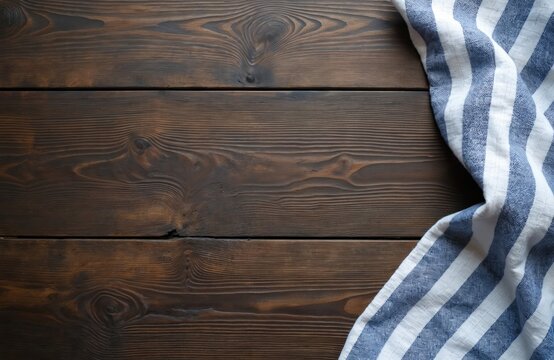Dark wood table surface with folded blue and white striped linen towel placed in corner. Empty space for food presentation or text. Rustic kitchen background, top view.