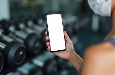 Woman wearing face mask holds phone with blank screen in gym near dumbbells. Fitness app mockup on smartphone screen. Healthy lifestyle digital device.