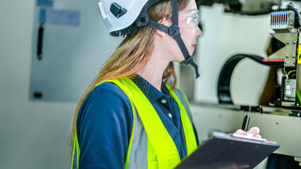 A female engineer in a hard hat and safety glasses inspects a high tech machine. She is holding a clipboard, doing a quality control check in a smart factory.