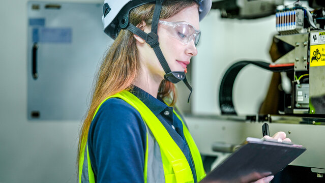 A female engineer in a hard hat and safety glasses inspects a high tech machine. She is holding a clipboard, doing a quality control check in a smart factory. - Powered by Adobe