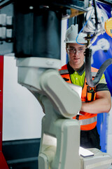 Fototapeta premium A male engineer in a hard hat holds a laptop while doing maintenance on a high tech industrial robotic arm. He is working in a futuristic smart factory.