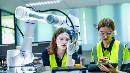 Two female robotics engineers in an R and D lab troubleshoot an AI cobot. One holds a tablet with diagnostic data while the other analyzes the robotic system.