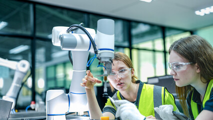 Two female engineers in safety vests collaborate in a tech lab. One woman in gloves inspects a high tech robotic arm while her colleague takes notes.